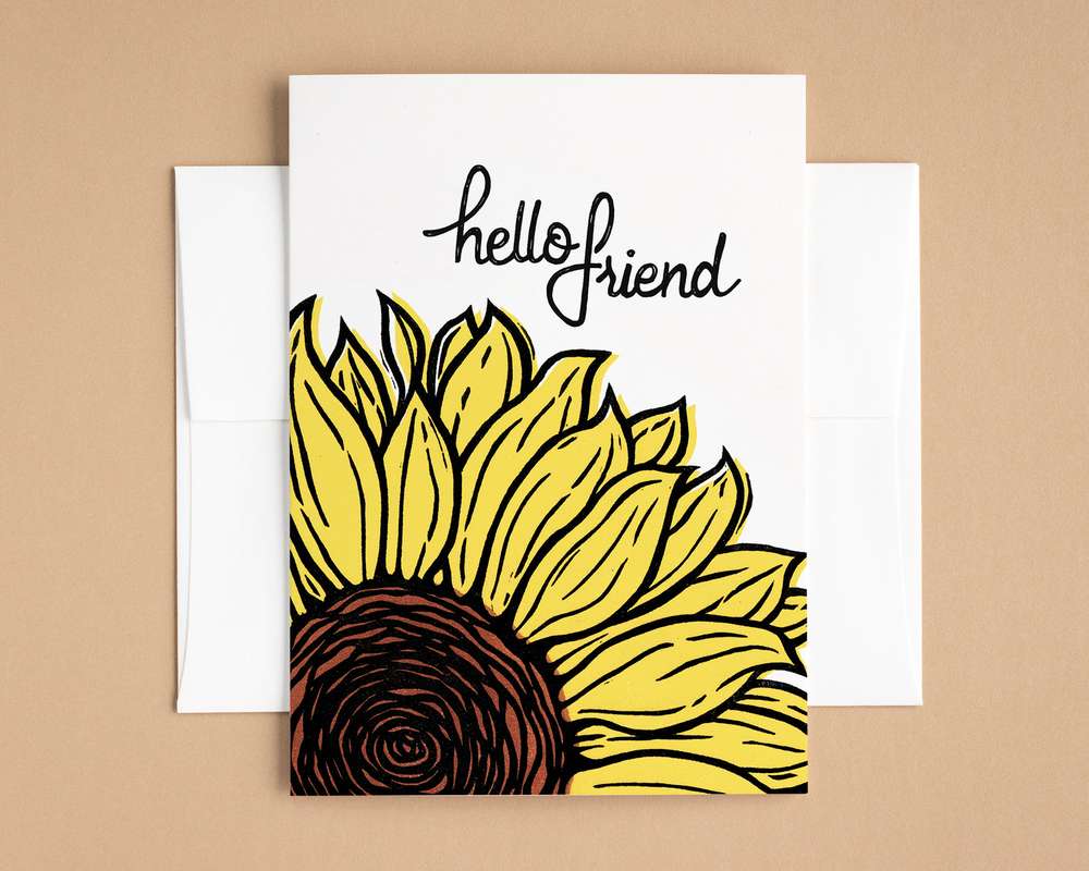 A vertical white card depicts a blockprinted yellow and black sunflower below text that reads 'Hello Friend'. The card sits on top of a white envelope, which lies on top of a brown backdrop.